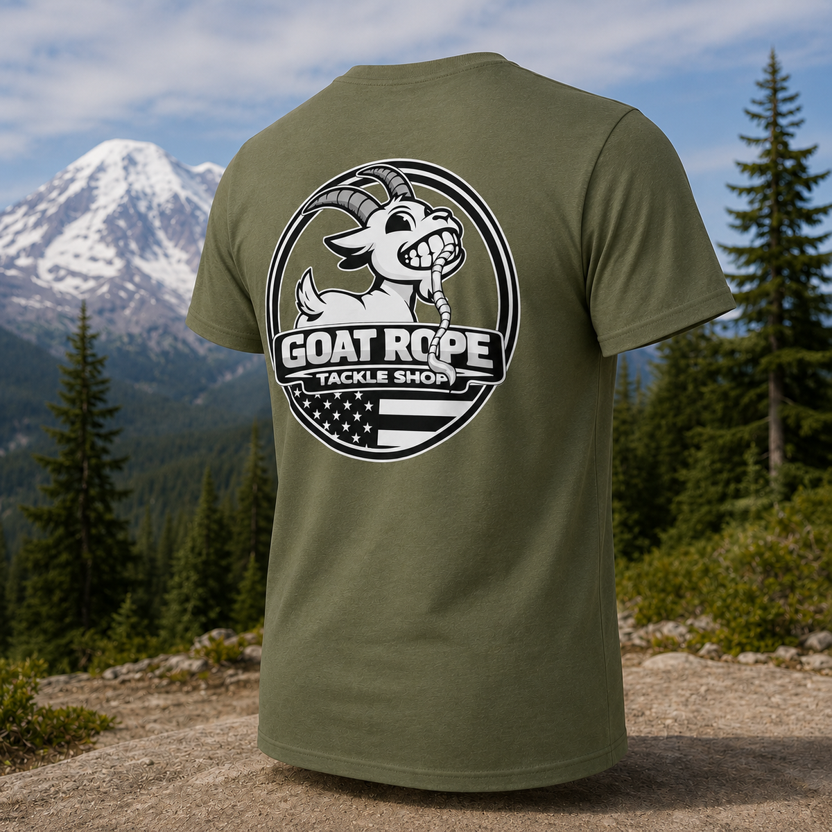 An olive green short-sleeve t-shirt is shown from the back, featuring a large circular graphic print of a cartoon goat and the text 'GOAT ROPE TACKLE SHOP' over an American flag motif. The shirt is displayed against a scenic outdoor background of a snowy mountain, pine trees, and a rocky path.