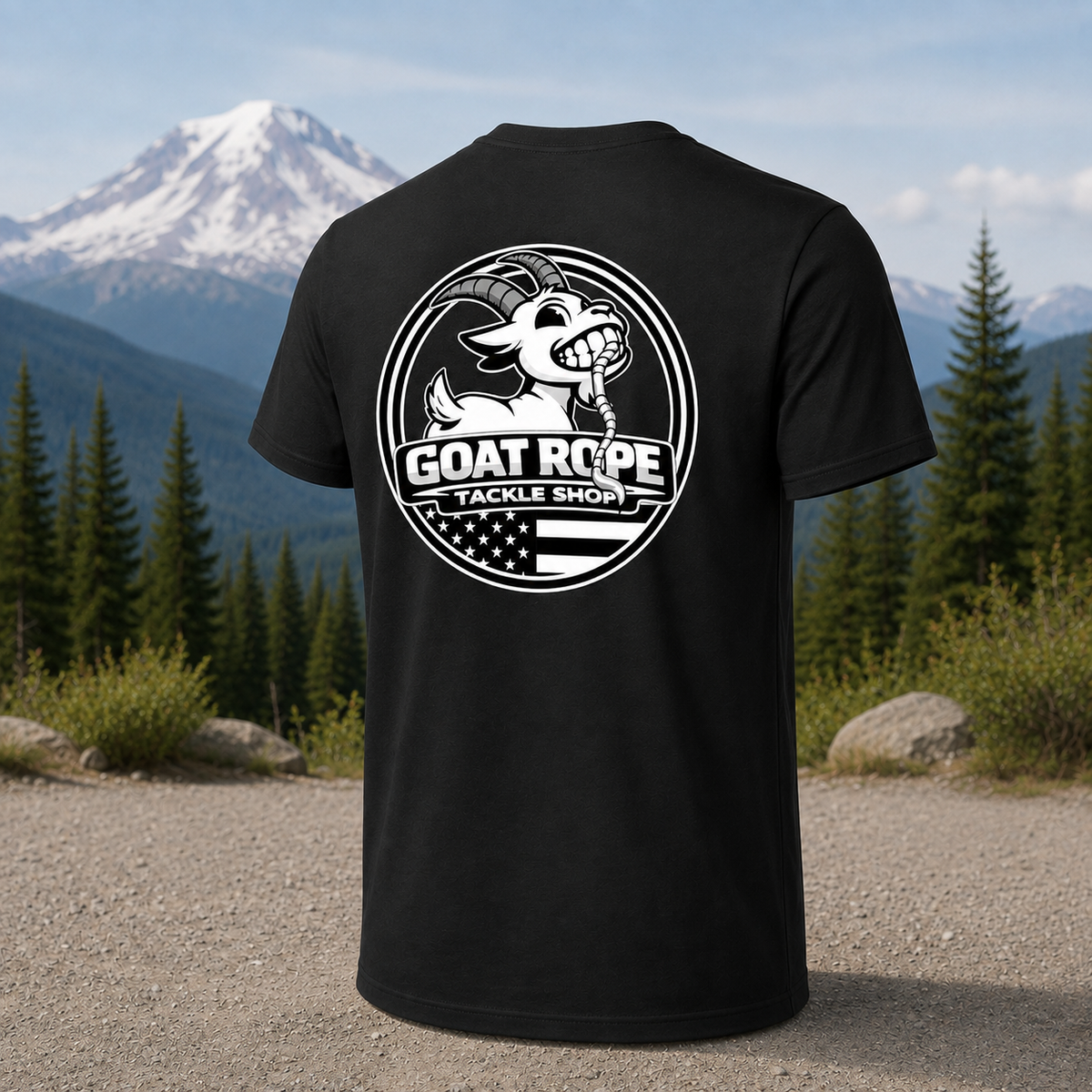 A black short-sleeve T-shirt shown from the back, featuring a large white circular graphic with a goat illustration and the text 'GOAT ROPE TACKLE SHOP.' The shirt is photographed outdoors in front of a mountain and pine forest, with no person present.