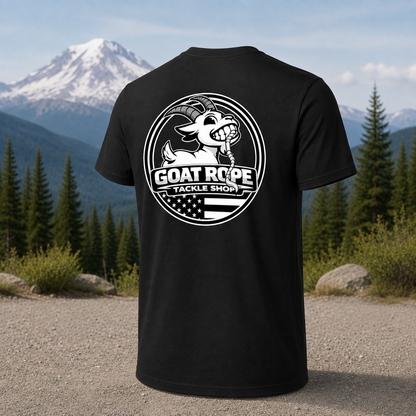 A black short-sleeve T-shirt shown from the back, featuring a large white circular graphic with a goat illustration and the text 'GOAT ROPE TACKLE SHOP.' The shirt is photographed outdoors in front of a mountain and pine forest, with no person present.