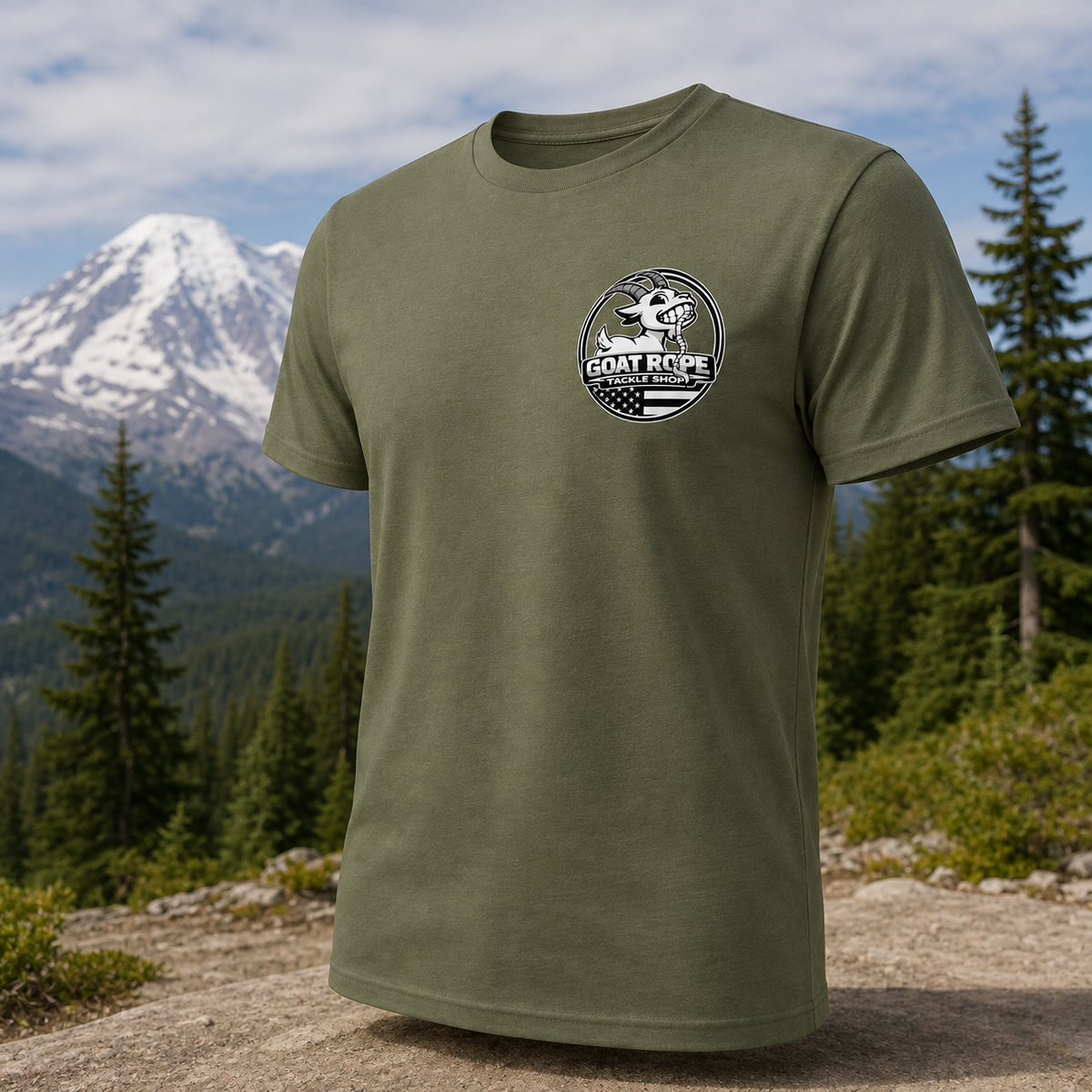 An olive green short-sleeve t-shirt with a circular 'GOAT ROPE' logo printed on the left chest, displayed against a blurred background of a mountain and pine forest.
