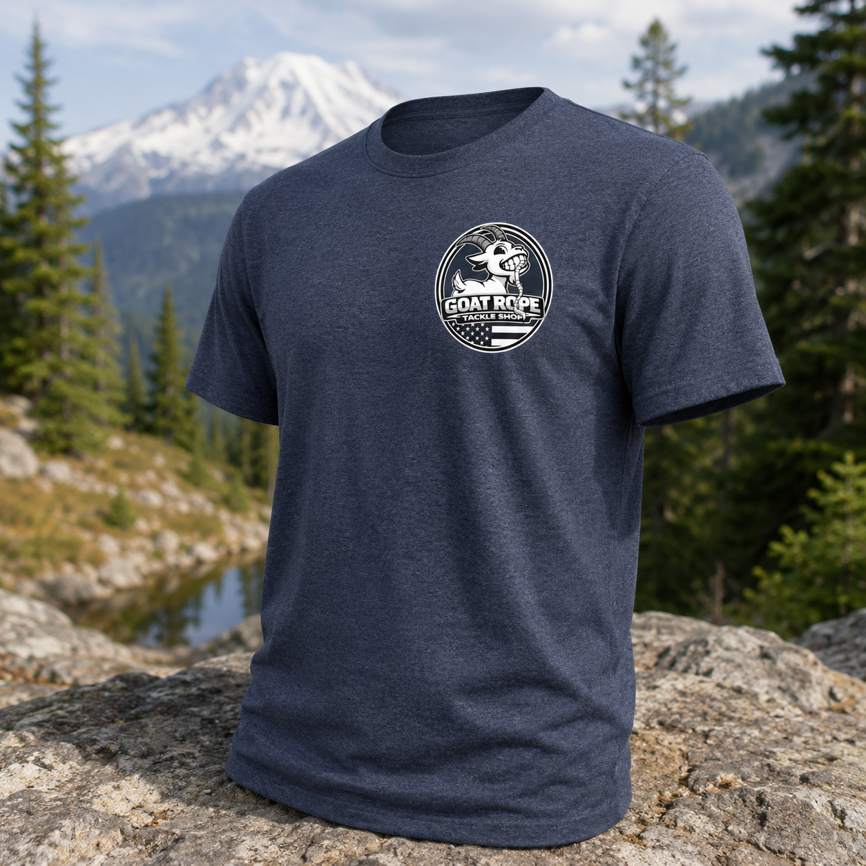 A heather navy blue short-sleeve t-shirt with a white circular 'Goat Rope' brand graphic on the left chest, displayed on a large rock with a mountain and forest background.