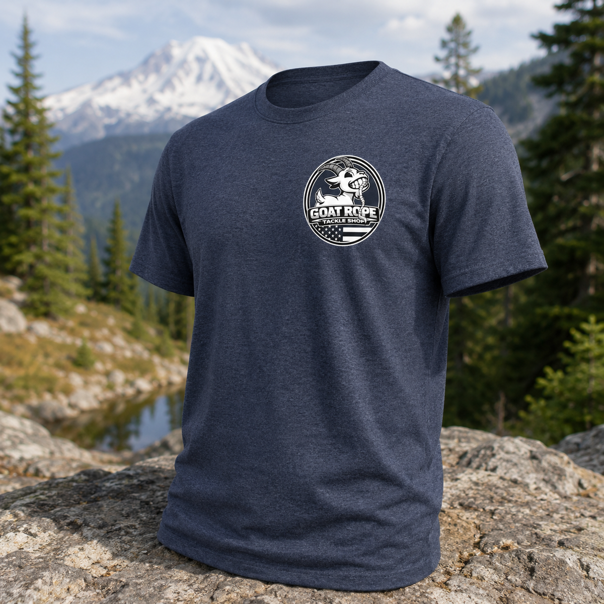 A heather navy blue short-sleeve t-shirt with a white circular 'Goat Rope' brand graphic on the left chest, displayed on a large rock with a mountain and forest background.