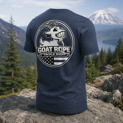 A navy blue heather short-sleeve t-shirt is shown from the back, featuring a large circular white graphic of a smiling goat mascot with the text 'GOAT ROPE TACKLE SHOP' and an American flag motif. The shirt is displayed outdoors on a large rock with a mountain, lake, and pine forest in the background.
