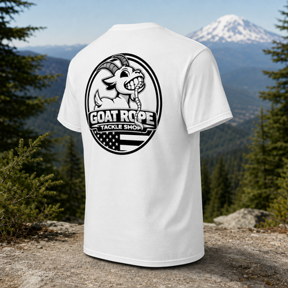 A white short-sleeve T-shirt shown from the back, featuring a large black circular graphic with a goat illustration and the text 'GOAT ROPE TACKLE SHOP.' The shirt is photographed outdoors on a rock with a mountain and pine forest in the background.