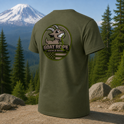 An olive green short-sleeve t-shirt shown from the back, featuring a large circular graphic of a cartoon goat with a green tongue sticking out and the text 'GOAT ROPE TACKLE SHOP'. The shirt is displayed outdoors against a background of pine trees, mountains, and rocks.