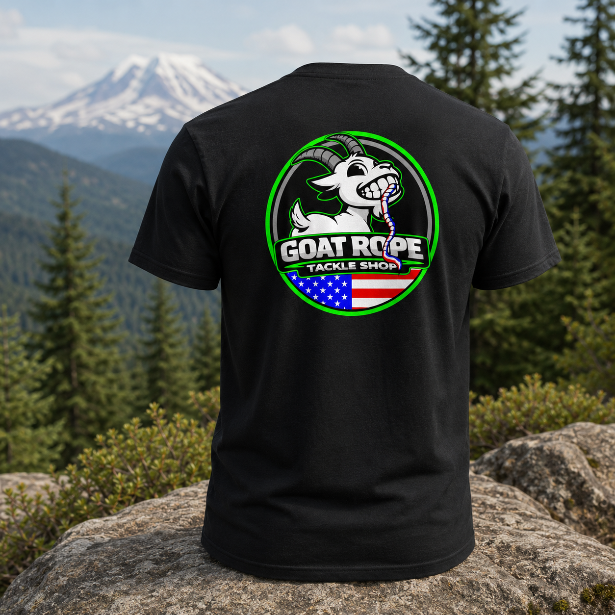 A black short-sleeve t-shirt shown from the back, featuring a large circular graphic with a white goat, green border, and the text 'GOAT ROPE TACKLE SHOP' over an American flag motif. The shirt is displayed on a large rock with a mountain and pine forest background.