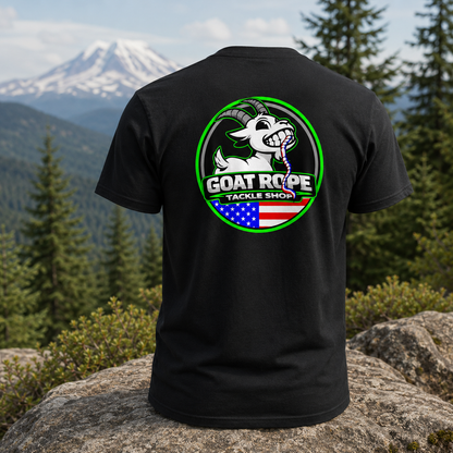 A black short-sleeve t-shirt shown from the back, featuring a large circular graphic with a white goat, green border, and the text 'GOAT ROPE TACKLE SHOP' over an American flag motif. The shirt is displayed on a large rock with a mountain and pine forest background.