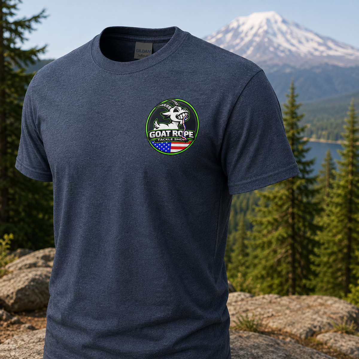 A heather navy blue short-sleeve t-shirt with a circular green, white, and red 'GOAT ROPE' logo on the left chest, displayed against a blurred mountain and forest background.