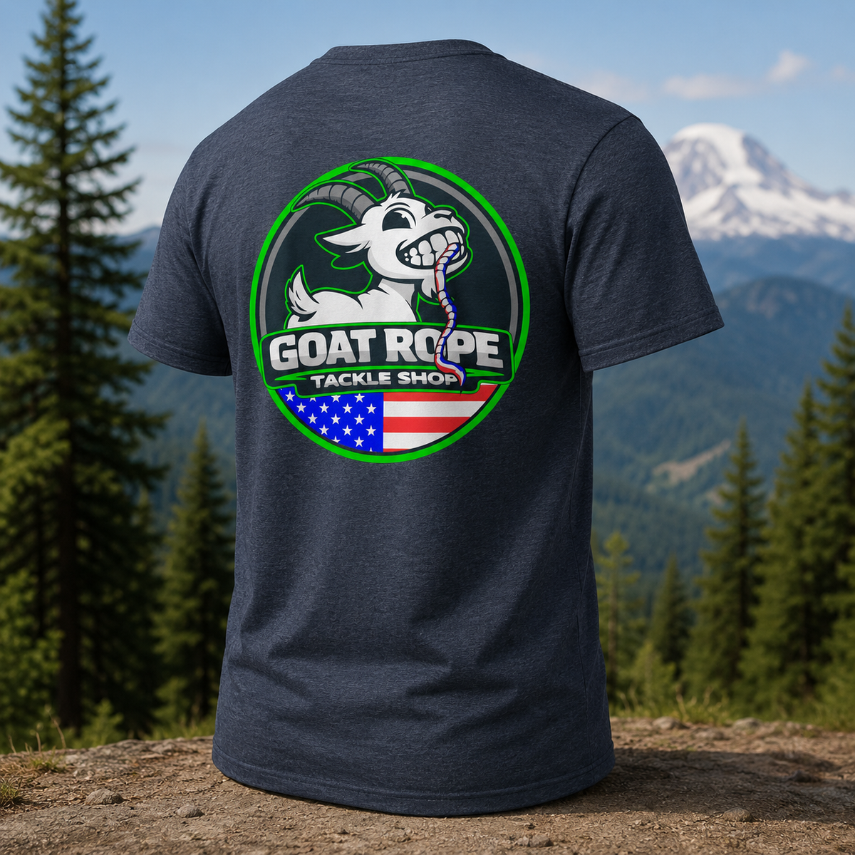 A dark heather gray t-shirt shown from the back, featuring a large circular graphic for 'GOAT ROPE TACKLE SHOP' with a white goat, green border, and an American flag motif. The shirt is displayed against a blurred outdoor mountain and pine forest background.