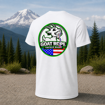 A white short-sleeve T-shirt shown from the back, featuring a large circular 'GOAT ROPE TACKLE SHOP' graphic with a cartoon goat, green border, and an American flag motif. The shirt is photographed outdoors against a mountain and pine tree background.