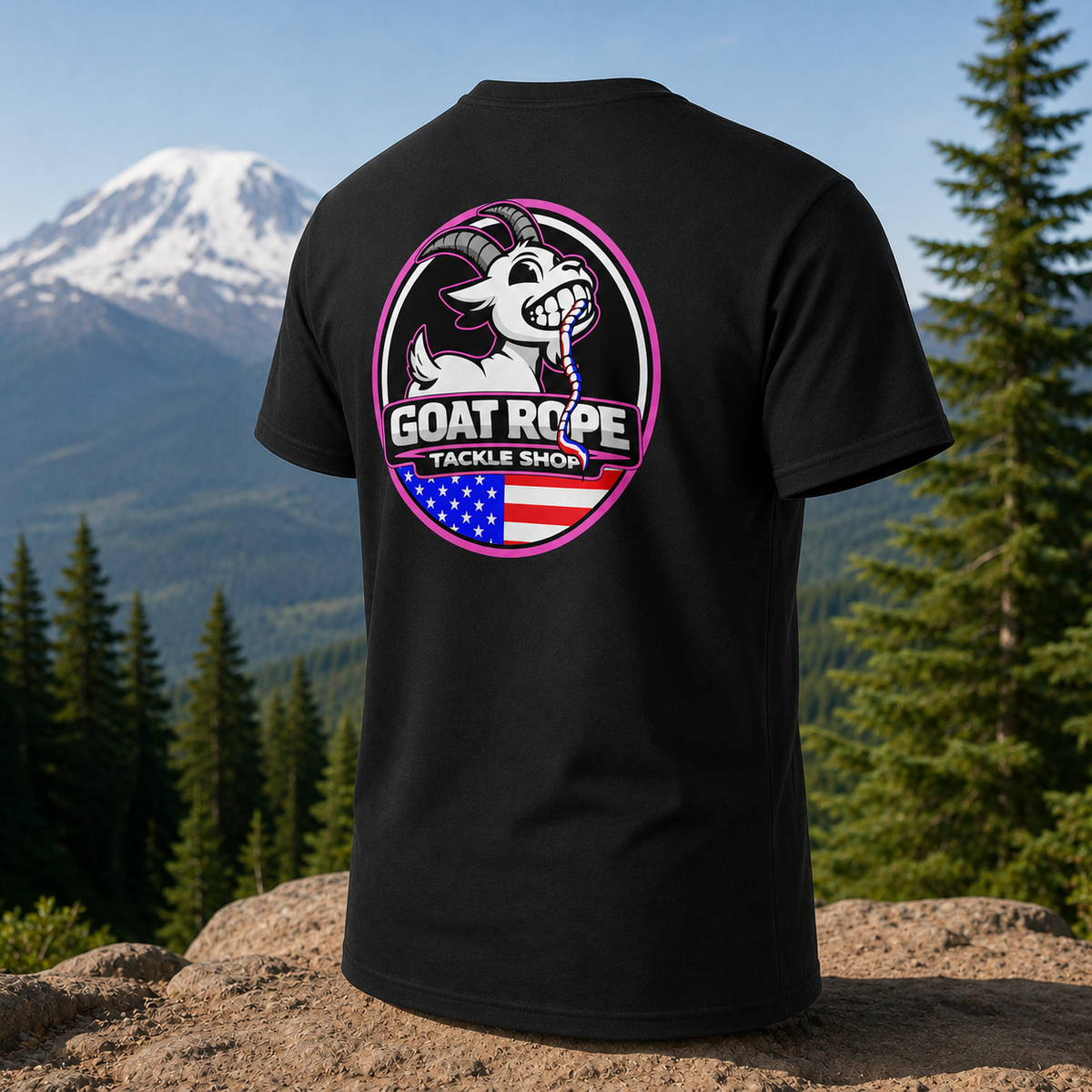 A black short-sleeve T-shirt shown from the back, featuring a large circular graphic with a white goat, pink outline, and the text 'GOAT ROPE TACKLE SHOP' over an American flag motif. The shirt is photographed outdoors on a rock with a mountain and pine forest in the background.