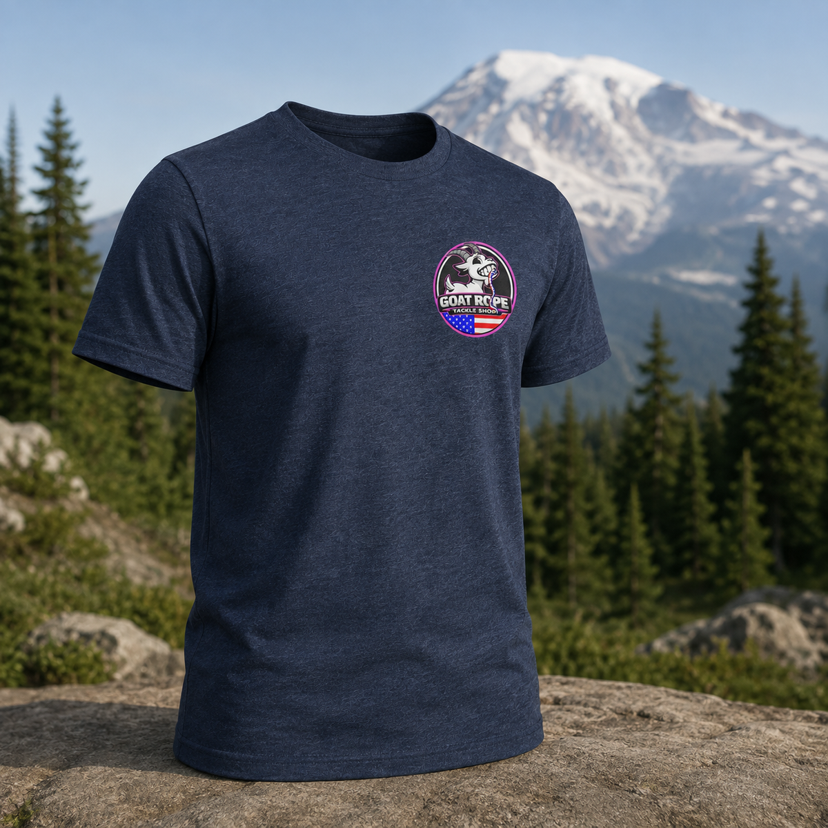 A navy blue heather short-sleeve t-shirt with a circular 'Goat Rope' brand graphic on the left chest, displayed on a rock with a mountain and pine forest background.