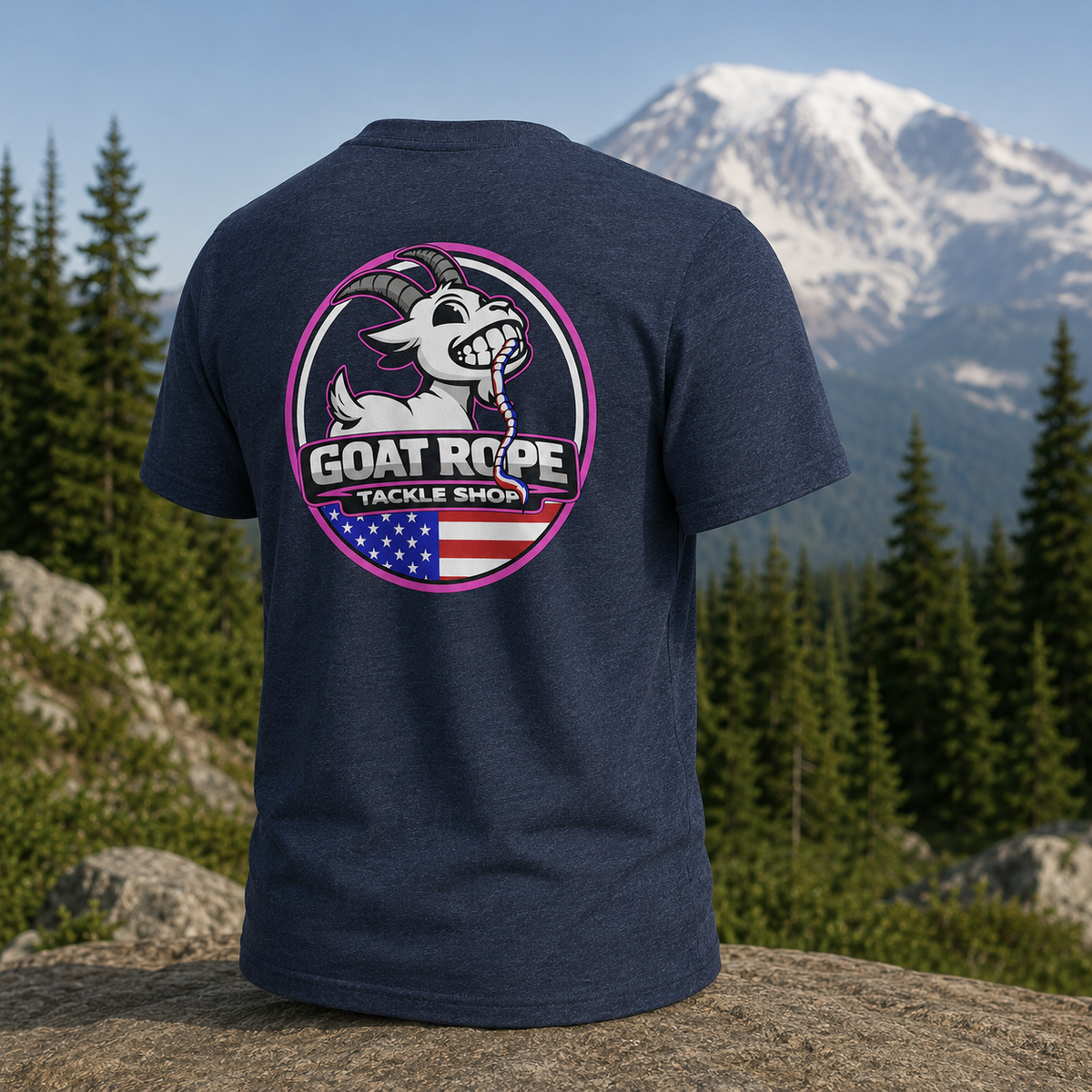 A heather navy blue t-shirt is shown from the back, featuring a large circular graphic of a cartoon goat with the text 'GOAT ROPE TACKLE SHOP' and an American flag motif. The shirt is displayed outdoors on a rock with a background of pine trees and a snow-capped mountain.