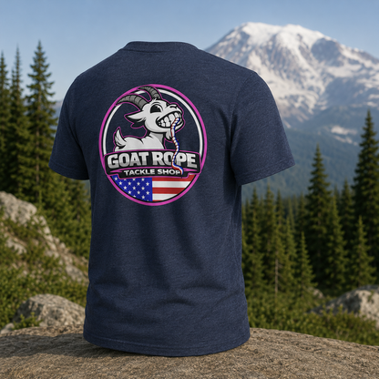 A heather navy blue t-shirt is shown from the back, featuring a large circular graphic of a cartoon goat with the text 'GOAT ROPE TACKLE SHOP' and an American flag motif. The shirt is displayed outdoors on a rock with a background of pine trees and a snow-capped mountain.