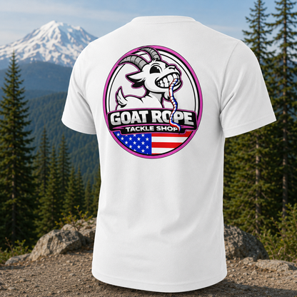 A white short-sleeve t-shirt shown from the back, featuring a large circular graphic with a cartoon goat, the text 'GOAT ROPE TACKLE SHOP', and an American flag motif. The shirt is displayed against a scenic background of a snowy mountain and pine trees.