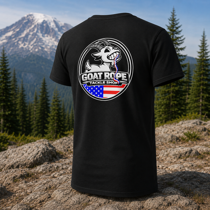 A black short-sleeve T-shirt is shown from the back, standing upright outdoors on a rocky mountain landscape with pine trees and a snow-capped peak in the background. The shirt features a large circular back graphic with a white goat holding a fishing line, the text 'GOAT ROPE TACKLE SHOP,' and an American flag motif in red, white, and blue.