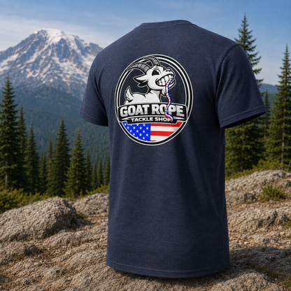 A navy blue short-sleeve t-shirt shown from the back, featuring a large circular graphic with a white goat, the text 'GOAT ROPE TACKLE SHOP', and an American flag motif. The shirt is displayed against a scenic outdoor background of a mountain and pine forest.