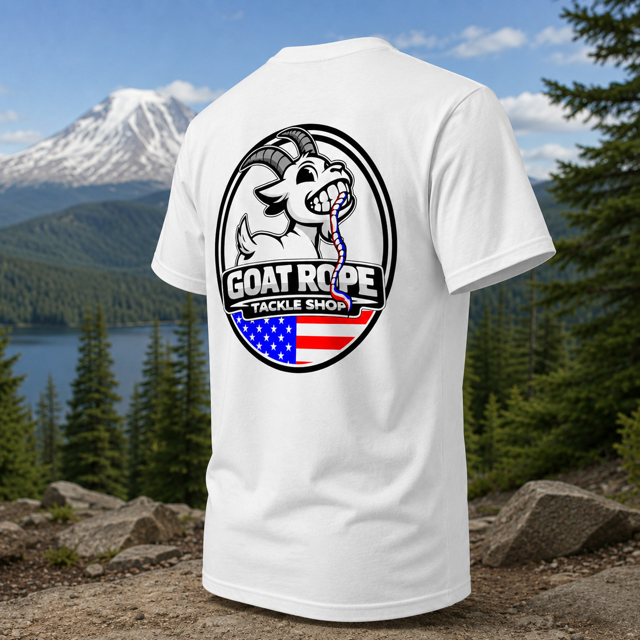 A white short-sleeve t-shirt with a large circular graphic on the back, featuring a cartoon goat and the text 'GOAT ROPE TACKLE SHOP' over an American flag design, shown against a mountain and lake background.