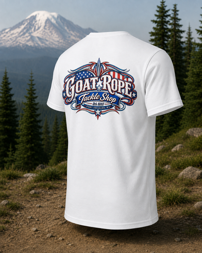 A white short-sleeve t-shirt is shown from the back, featuring a large patriotic graphic with the text 'GOAT ROPE Tackle Shop' in red, white, and blue. The shirt is displayed against a scenic background of a snowy mountain and pine trees.