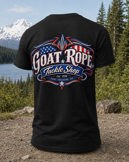 A black short-sleeve T-shirt shown from the back, featuring a large patriotic graphic that reads "GOAT ROPE Tackle Shop" with red, white, and blue flag-style details. The shirt is photographed outdoors against a scenic background of trees, water, rocks, and a mountain.