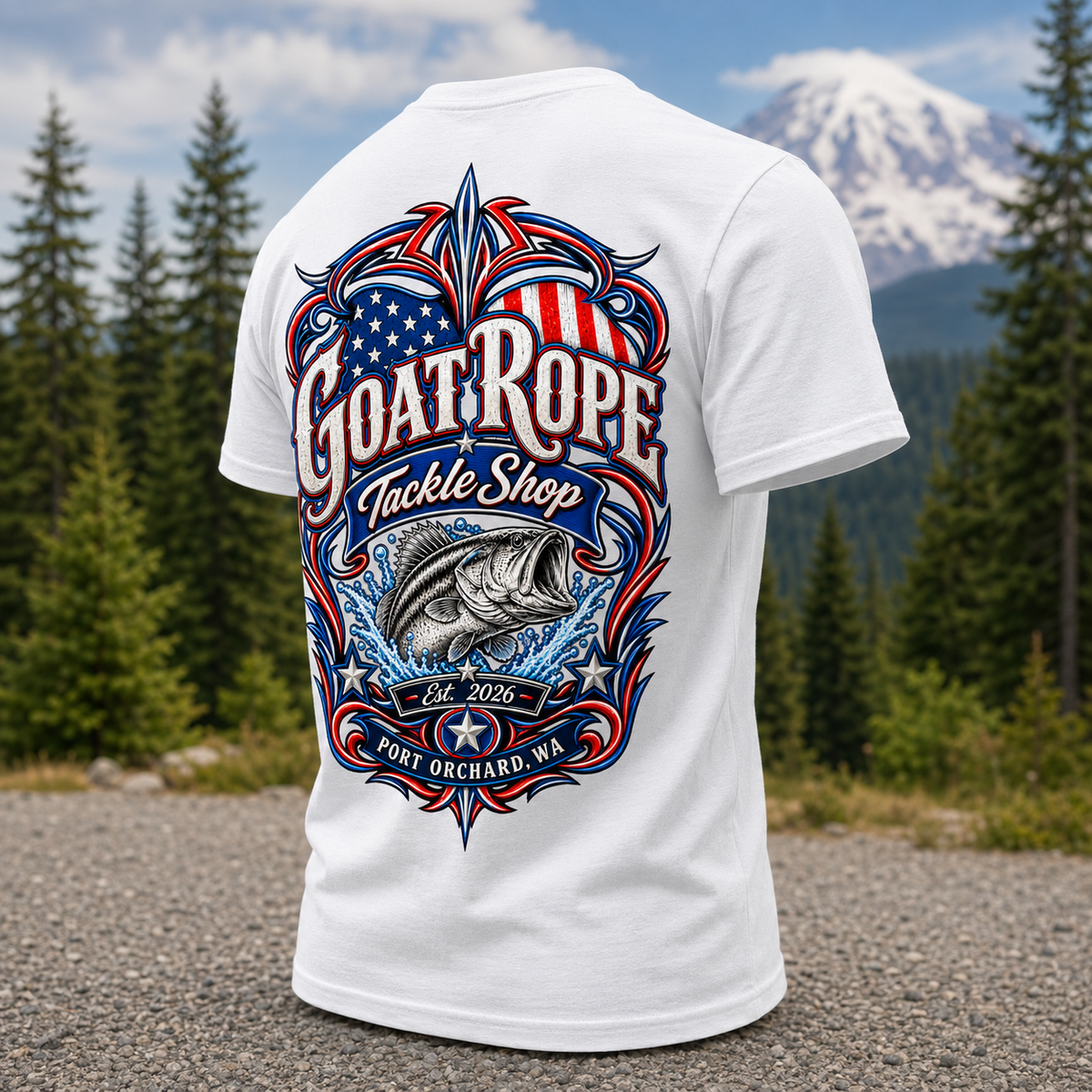 A white short-sleeve T-shirt shown from the back, featuring a large patriotic fishing graphic with the text 'Goat Rope Tackle Shop,' a fish illustration, and 'Port Orchard, WA.' The shirt is photographed outdoors on a gravel path with pine trees and a snow-capped mountain in the background.