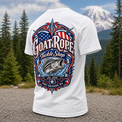A white short-sleeve T-shirt shown from the back, featuring a large patriotic fishing graphic with the text 'Goat Rope Tackle Shop,' a fish illustration, and 'Port Orchard, WA.' The shirt is photographed outdoors on a gravel path with pine trees and a snow-capped mountain in the background.