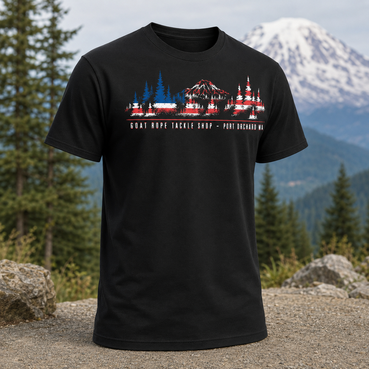 A black short-sleeve graphic t-shirt displayed against a scenic mountain and forest background. A black short-sleeve crewneck t-shirt featuring a graphic print of a mountain and fishing tackle shop in a stylized American flag pattern, with the text 'GOAT ROPE TACKLE SHOP - PORT Orchard WA' printed below.