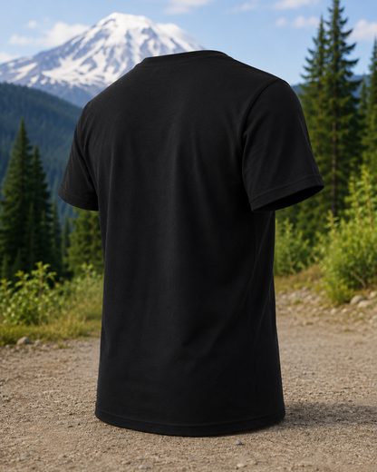 A black short-sleeve crewneck t-shirt shown from the back, positioned on a gravel path with a forest and a snow-capped mountain in the background.