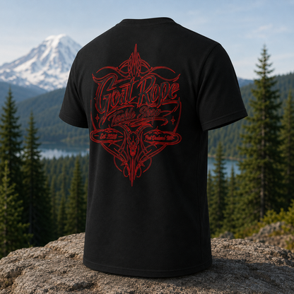 A black short-sleeve t-shirt shown from the back, featuring a large red graphic print with the text 'Goat Rope' and 'Tackle Shop' over a stylized horned emblem. The shirt is displayed on a rock with a blurred mountain and forest background.