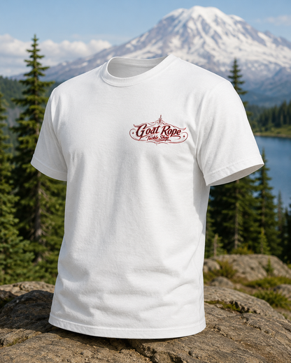 A white short-sleeve t-shirt with a maroon 'Goat Rope' logo on the left chest, displayed on a rock with a mountain and lake background.